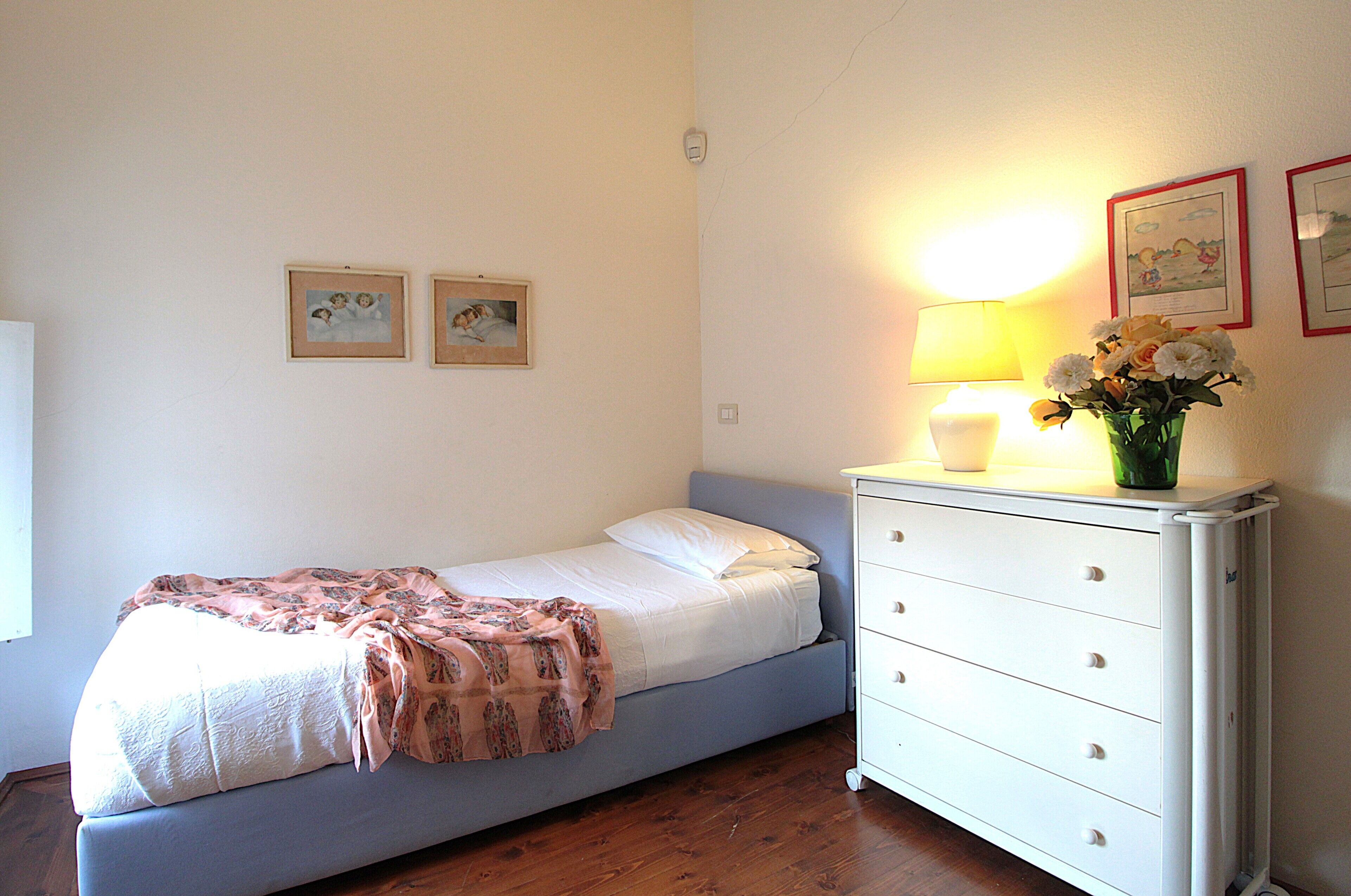 Habitación con una cama individual, suelo de madera, una mesita de noche con lámpara y dos cuadros en la pared.