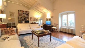 Un salón con un sofá blanco, una mesa de centro con libros, una estantería, un televisor y un ventanal con vistas.