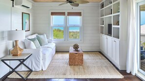 A room with a white sofa, a small table, a ceiling fan, and a view of the ocean through large windows. - New Luxury Beachfront Bungalows w/Heated Pool, Pink-Sand Beach (Governors Harbour)