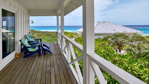 A wooden deck with two blue chairs and a small table, overlooking a beach with clear blue water and a white building in the distance. - New Luxury Beachfront Bungalows w/Heated Pool, Pink-Sand Beach (Governors Harbour)