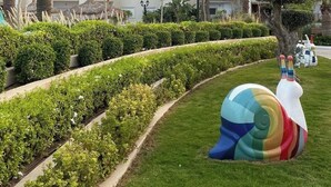 A landscaped garden with neatly trimmed hedges, a palm tree, and a large colorful sculpture. - Pool-view apartment in a charming, well-secured residence (Monastir)