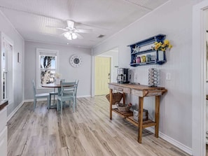 A kitchen with a dining table, chairs, a coffee maker, and a shelf with decorative items.