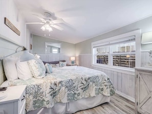 A bedroom with a bed, bedside tables, a nightstand, a ceiling fan, a window with blinds, and a wall-mounted mirror.