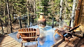 Une table en verre, deux chaises Adirondack en bois, une lanterne en métal et un paysage boisé en arrière-plan.
