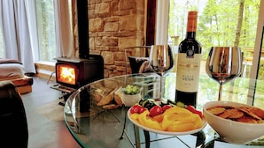 Une pièce chaleureuse avec une cheminée en pierre, une table en verre où sont disposés de la nourriture et du vin, et une vue sur la verdure environnante.
