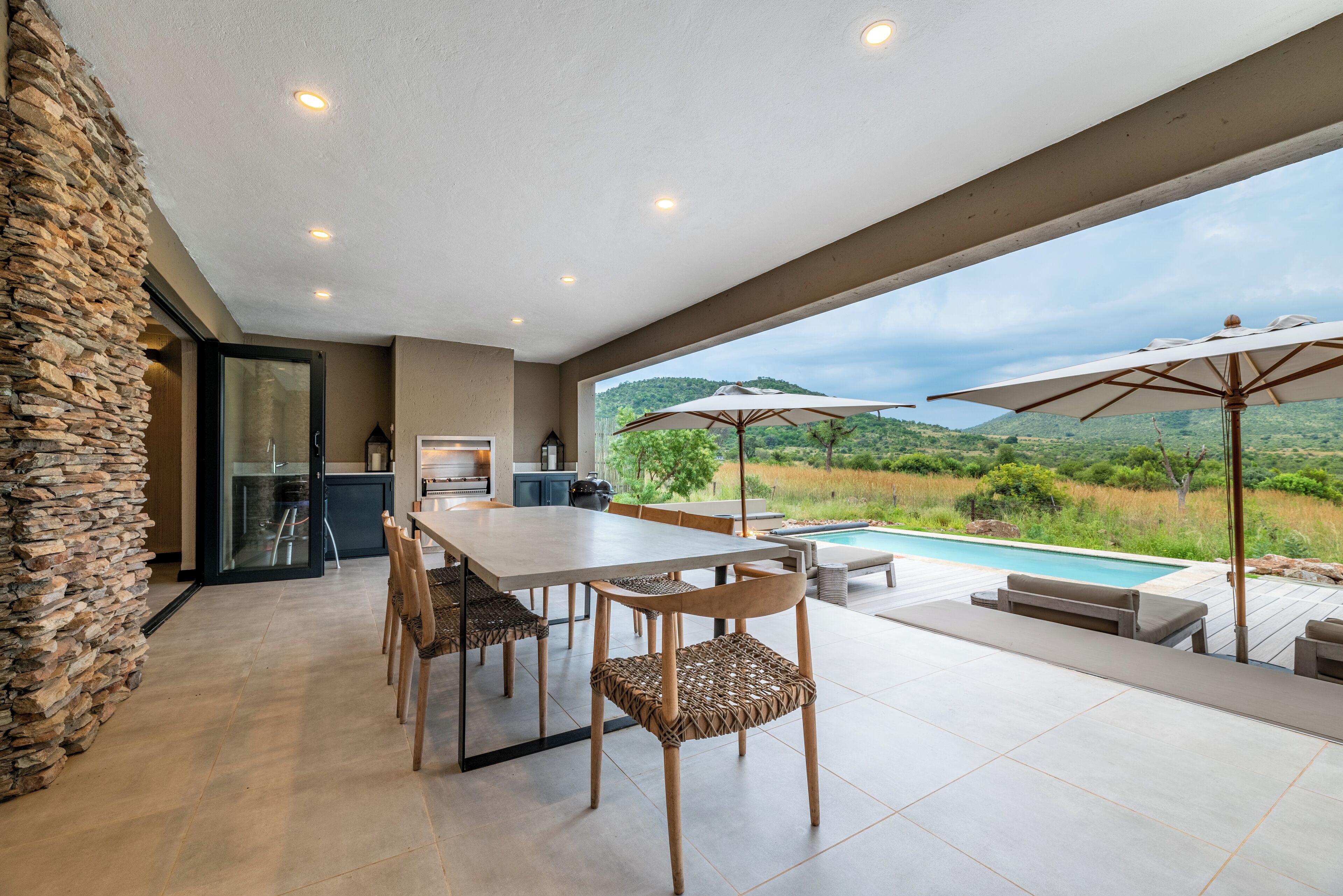 A modern outdoor dining area with a stone wall, a pool, and a view of the countryside.