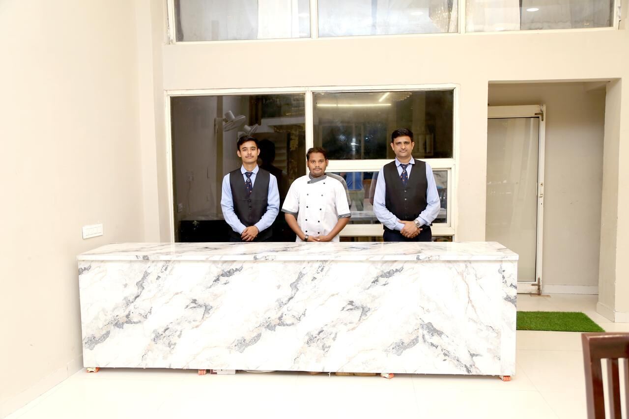 Three individuals standing behind a marble reception desk in a room with large windows and a green carpet.