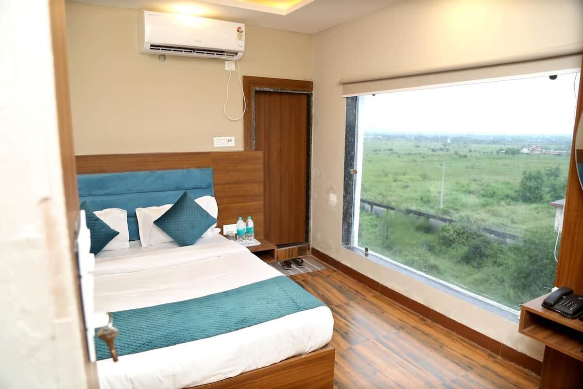 Quarto de hotel com uma cama grande, cabeceira de madeira, janela com vista para o verde, ar condicionado e mesa de cabeceira com telefone e garrafas de água.