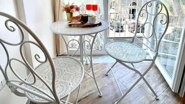 A balcony with two metal chairs, a small round table with a vase and red glasses, and a view of buildings through a window with white curtains. - GRANORO B&B (Gragnano)