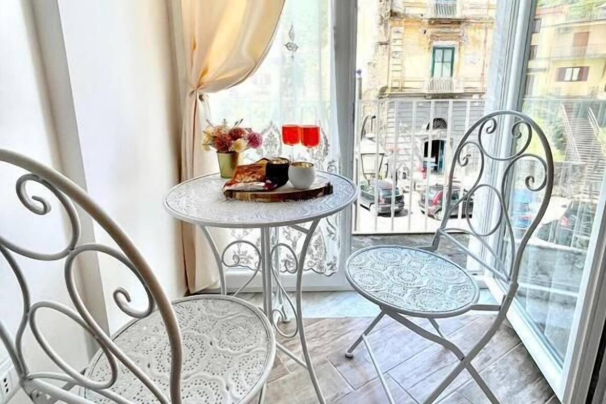 A balcony with two metal chairs, a small round table with a vase and red glasses, and a view of buildings through a window with white curtains.