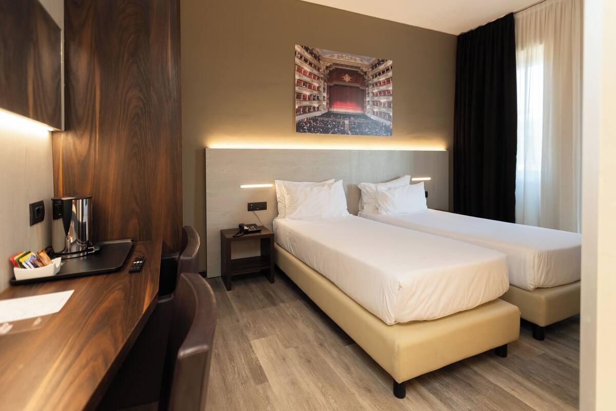 Habitación de hotel con dos camas, un escritorio con lámpara, una mesita con teléfono y un cuadro colgado en la pared de un auditorio.