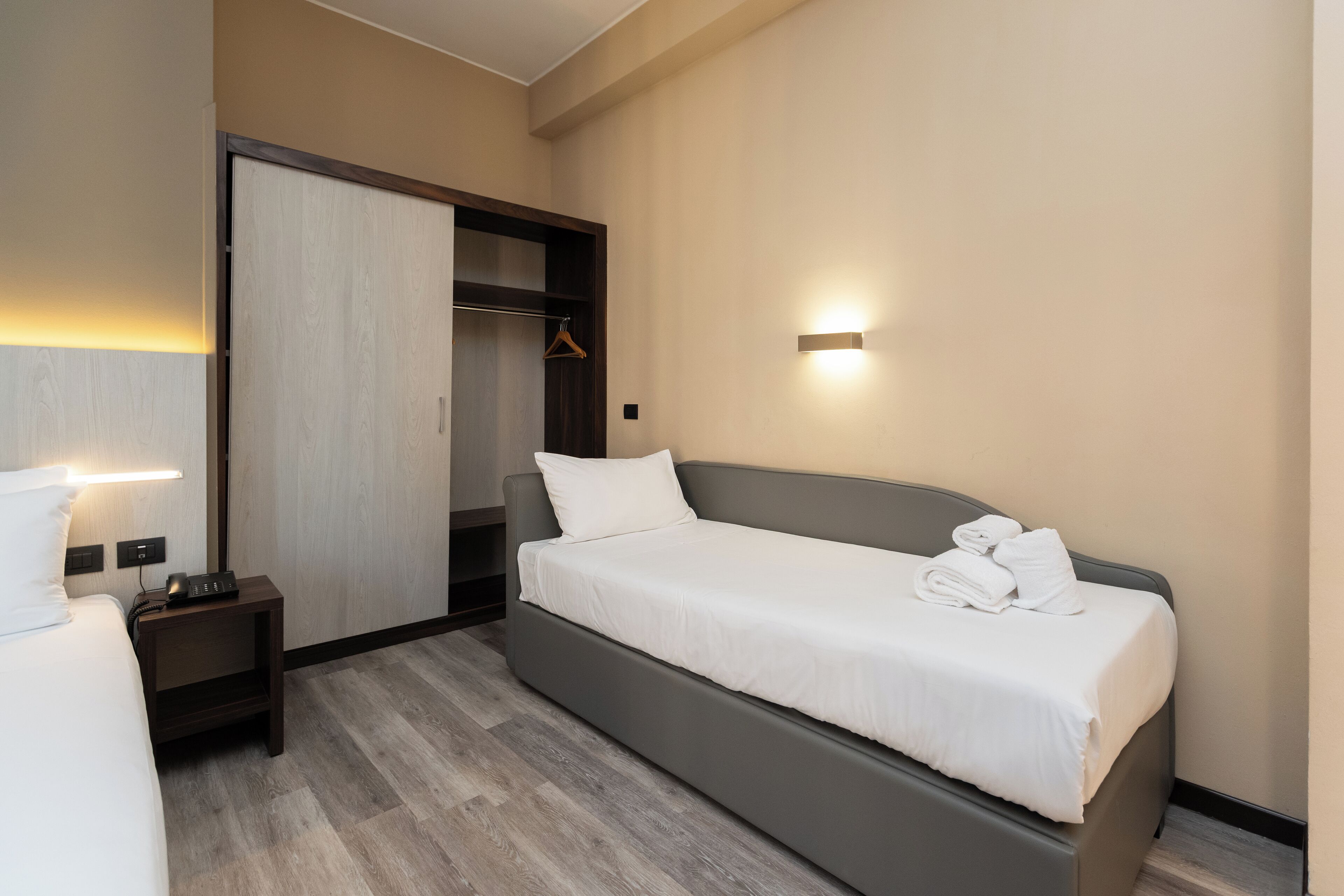 Camera d'albergo moderna con un letto singolo, un comodino con lampada, un armadio e una luce a parete.