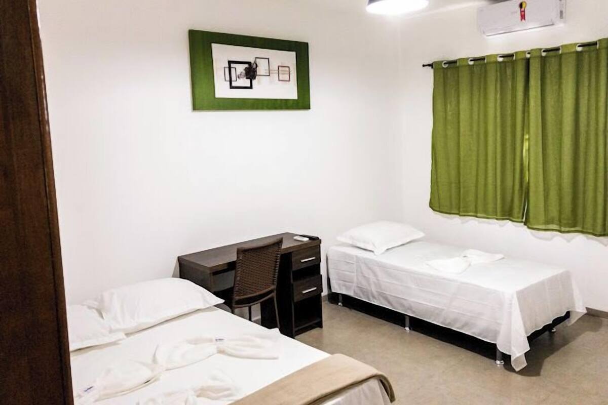 A small, clean room with two beds, a desk, a chair, and a green curtain.