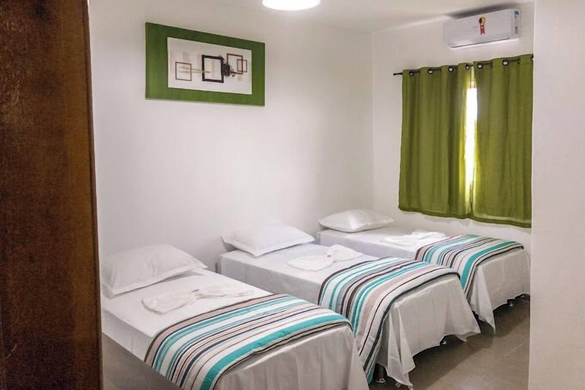 A room with three single beds, each with white linens and striped blankets. There is a ceiling light fixture and a wall-mounted air conditioning unit.