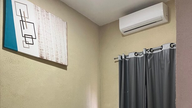 A single bed with a white mattress and a white pillow, a wall-mounted air conditioner, and a window with grey curtains. - Hotel JEM (Itiquira)