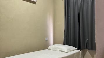 A single bed with a white mattress and a white pillow, a wall-mounted air conditioner, and a window with grey curtains.