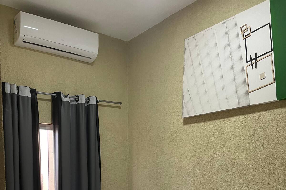 A single bed with white bedding in a room with beige walls, an air conditioning unit above the bed, and a window with dark curtains.