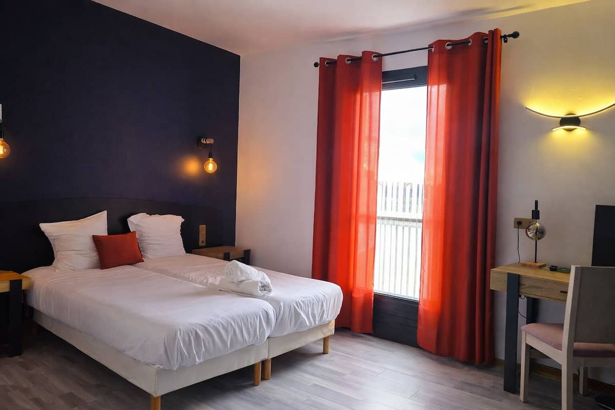 Un dormitorio con una cama, dos mesitas de noche, un escritorio con una silla y una ventana con cortinas rojas.