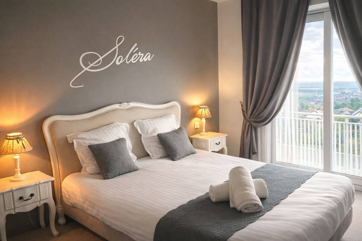 Una cama bien tendida con sábanas blancas y almohadas grises, una lámpara de noche y una pared con la palabra 'Solera' en cursiva.
