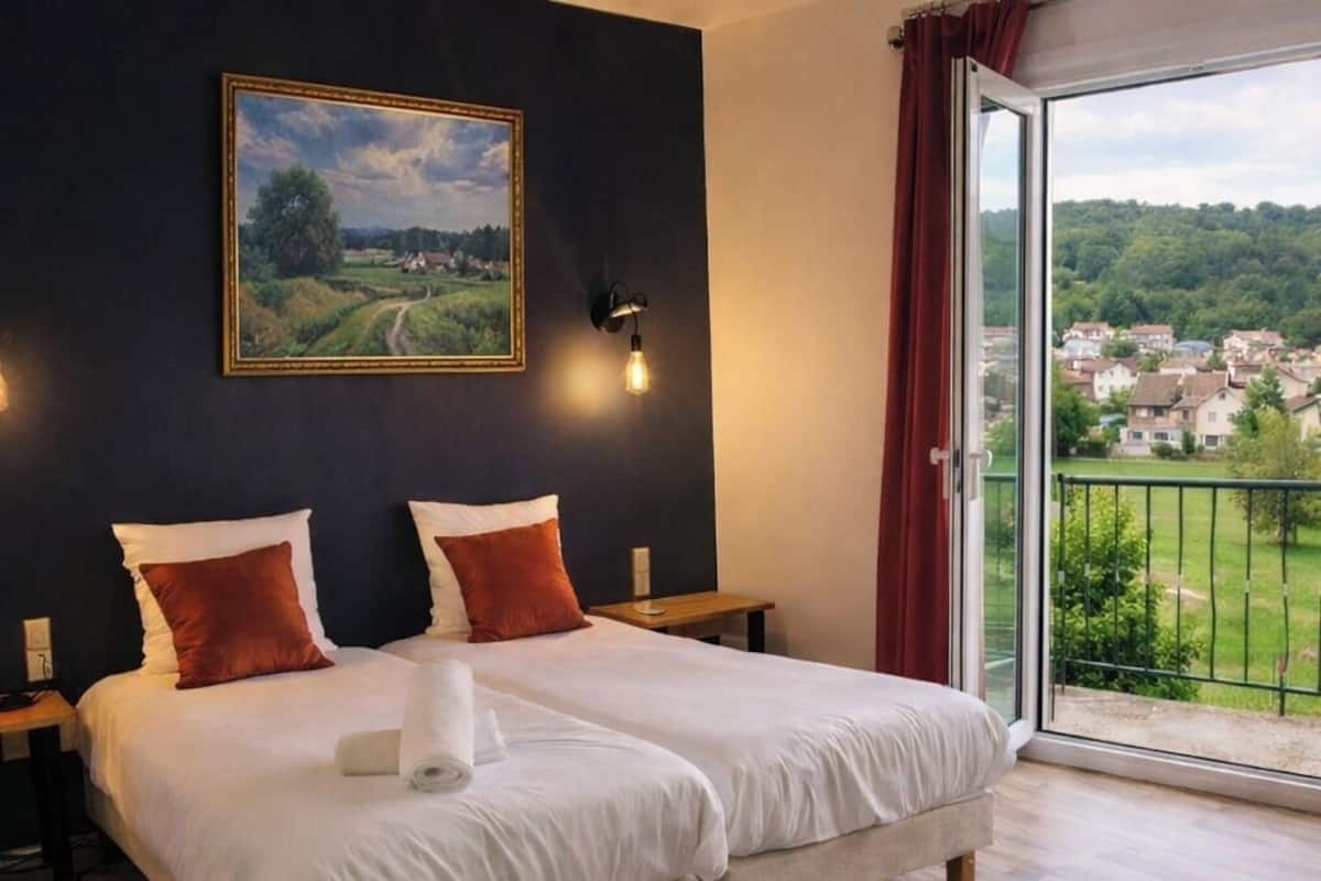 Cama doble con sábanas blancas y dos almohadas, una mesita de noche, una lámpara de pared y un ventanal con vistas a un paisaje verde.