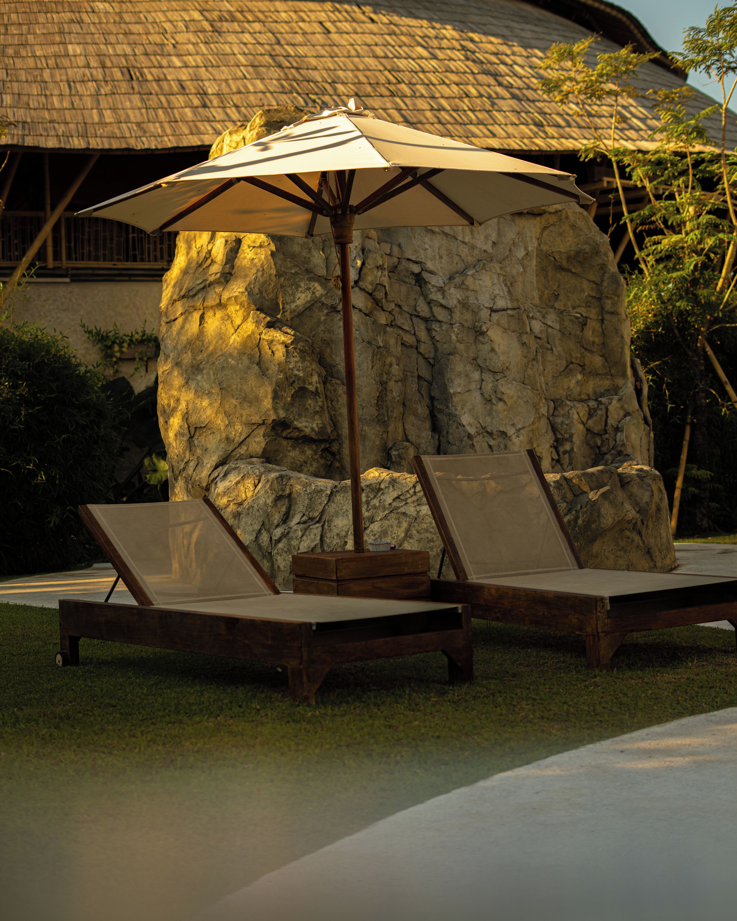 A lounge area with two sun loungers and a large umbrella, set against a backdrop of a stone wall and a thatched roof structure.