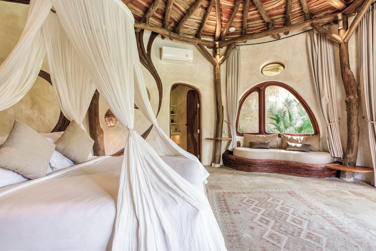 A bedroom with a canopy bed, a built-in bench, and a view through a window.