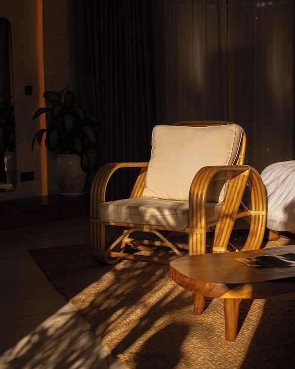 A wicker chair with a cushion, a small wooden table, and a potted plant in a room with a dark curtain background. - Nuanu Suites & Accommodations (Tabanan)