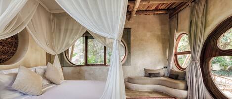 A bedroom with a canopy bed, a red and beige patterned rug, and a view of greenery through large windows.