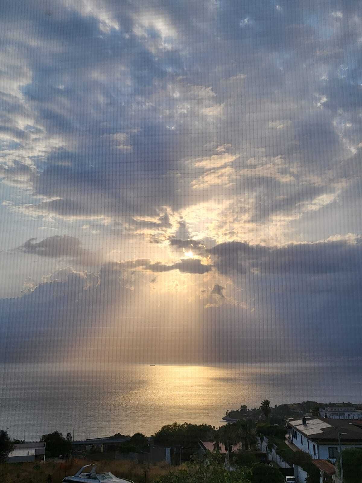 A coastal view with a sunset over the water, houses in the foreground, and a cloudy sky.