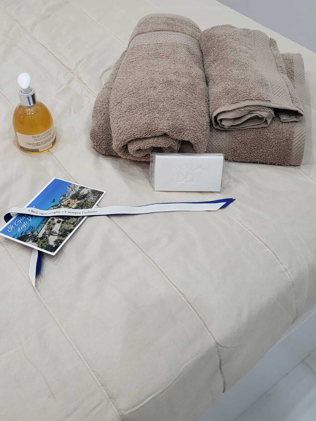 A neatly made bed with folded towels, a bottle of Bergamone, a card, and a white box on a white bedspread.