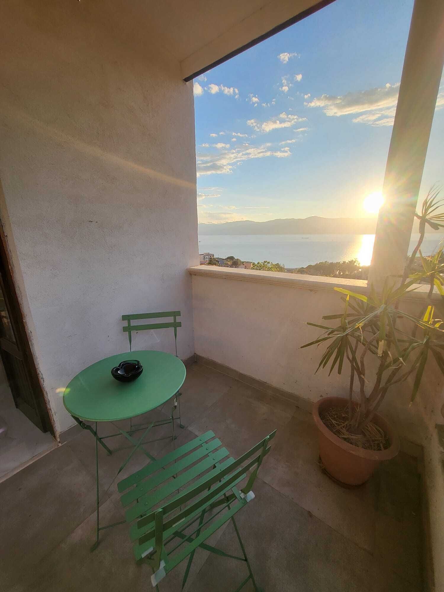 Un balcón con una mesa y sillas verdes, una planta en maceta y vistas al mar y a las montañas durante el atardecer.