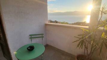 Ein Balkon mit einem grünen Tisch und Stühlen, einem Topfpflanzen und Blick auf das Meer und die Berge bei Sonnenuntergang.