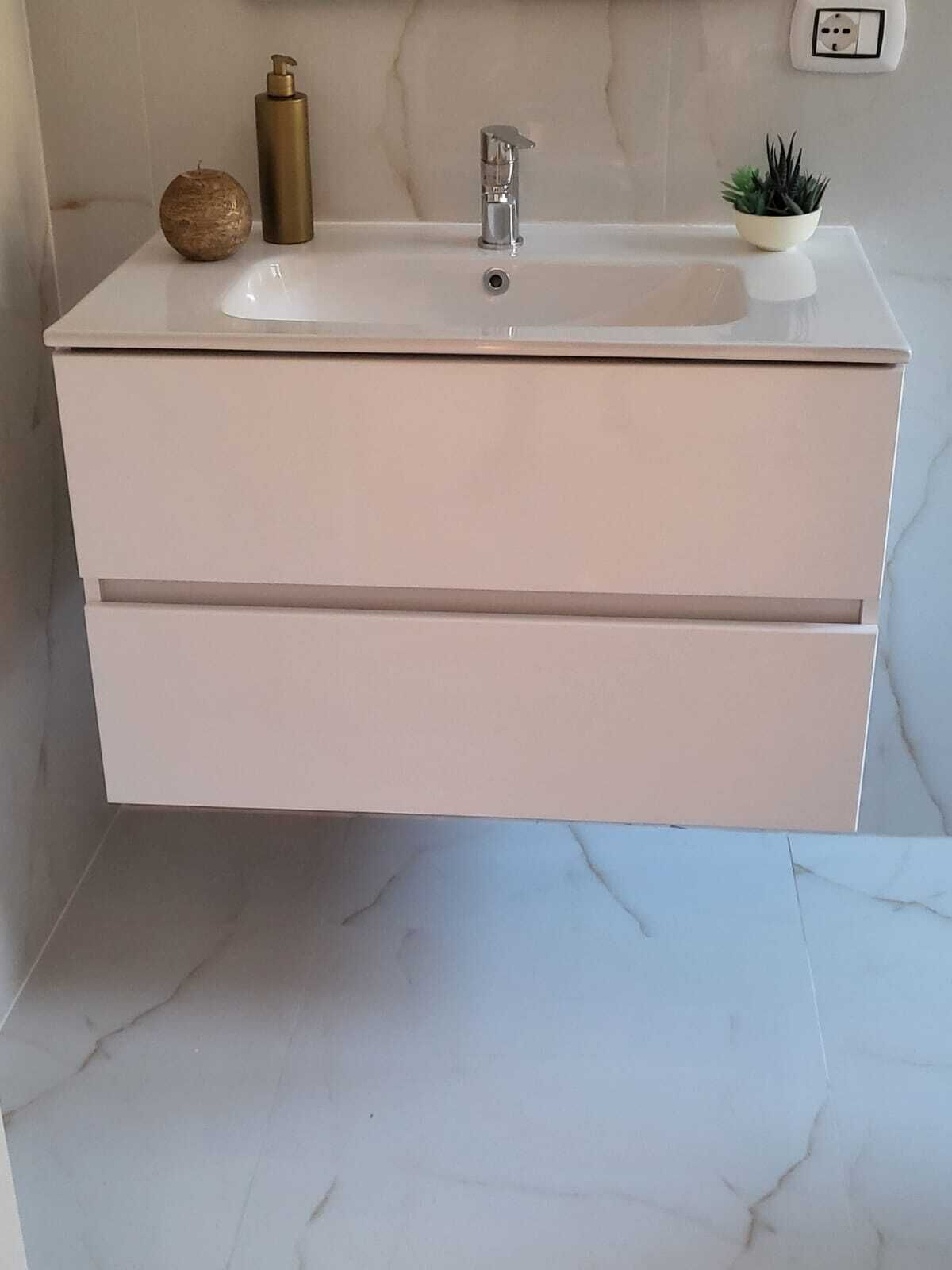 A white bathroom vanity with a sink, two drawers, a soap dispenser, a small plant, and a wall socket.