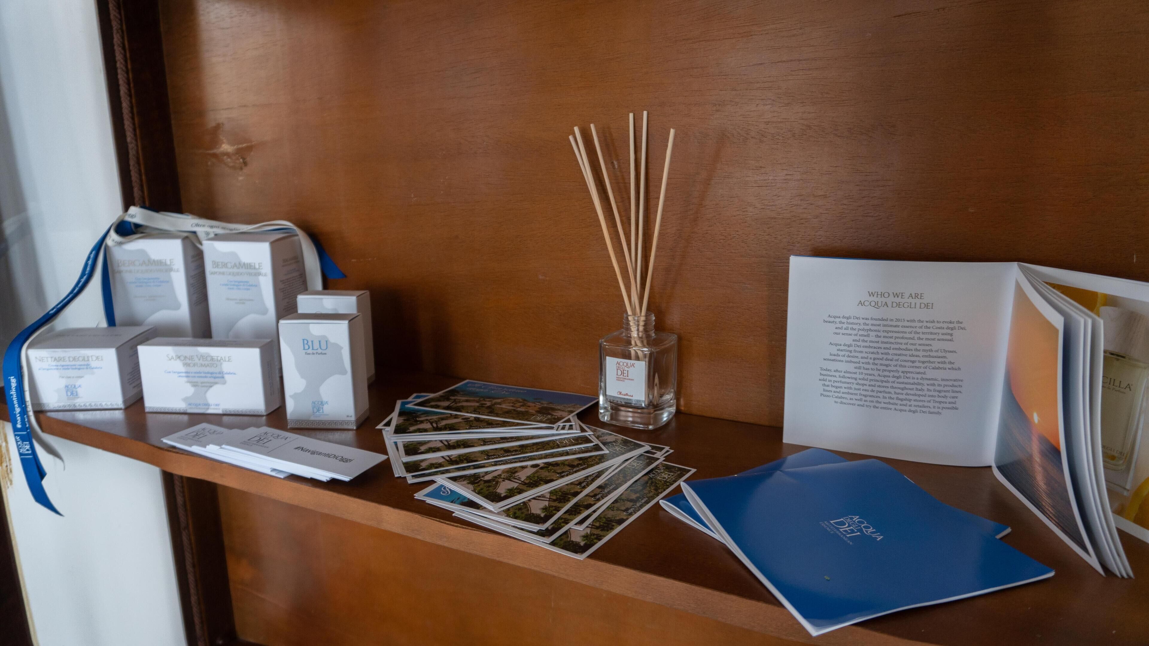 Una reception in legno con diversi oggetti, tra cui scatole etichettate 'Acqua Dei', un diffusore con bastoncini di giunchi e brochure.