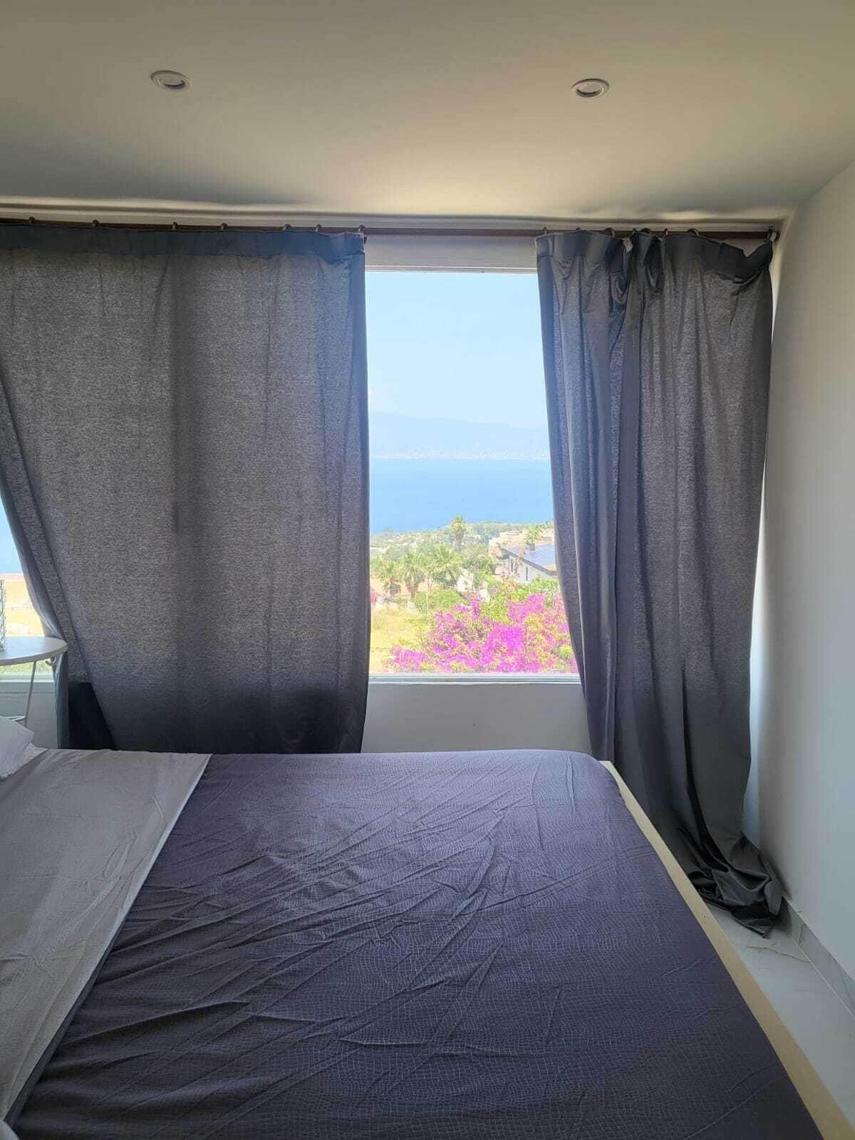 Una camera da letto con un letto, una finestra con tende e vista sul mare e sul verde.