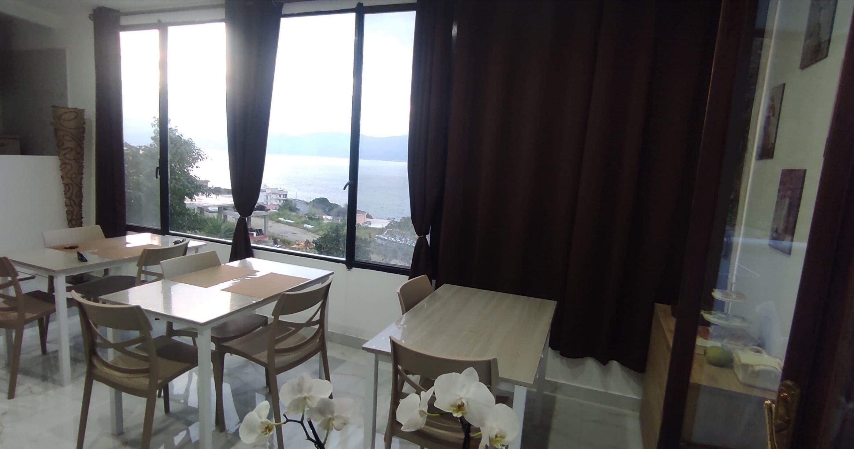 A dining area with a view of the sea, featuring a table and chairs, and a vase with white flowers.