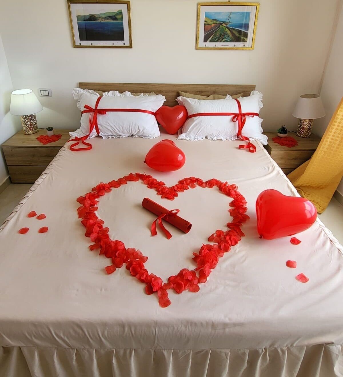 A bed with a heart-shaped arrangement of red flowers and balloons, white pillows, and a wooden headboard.