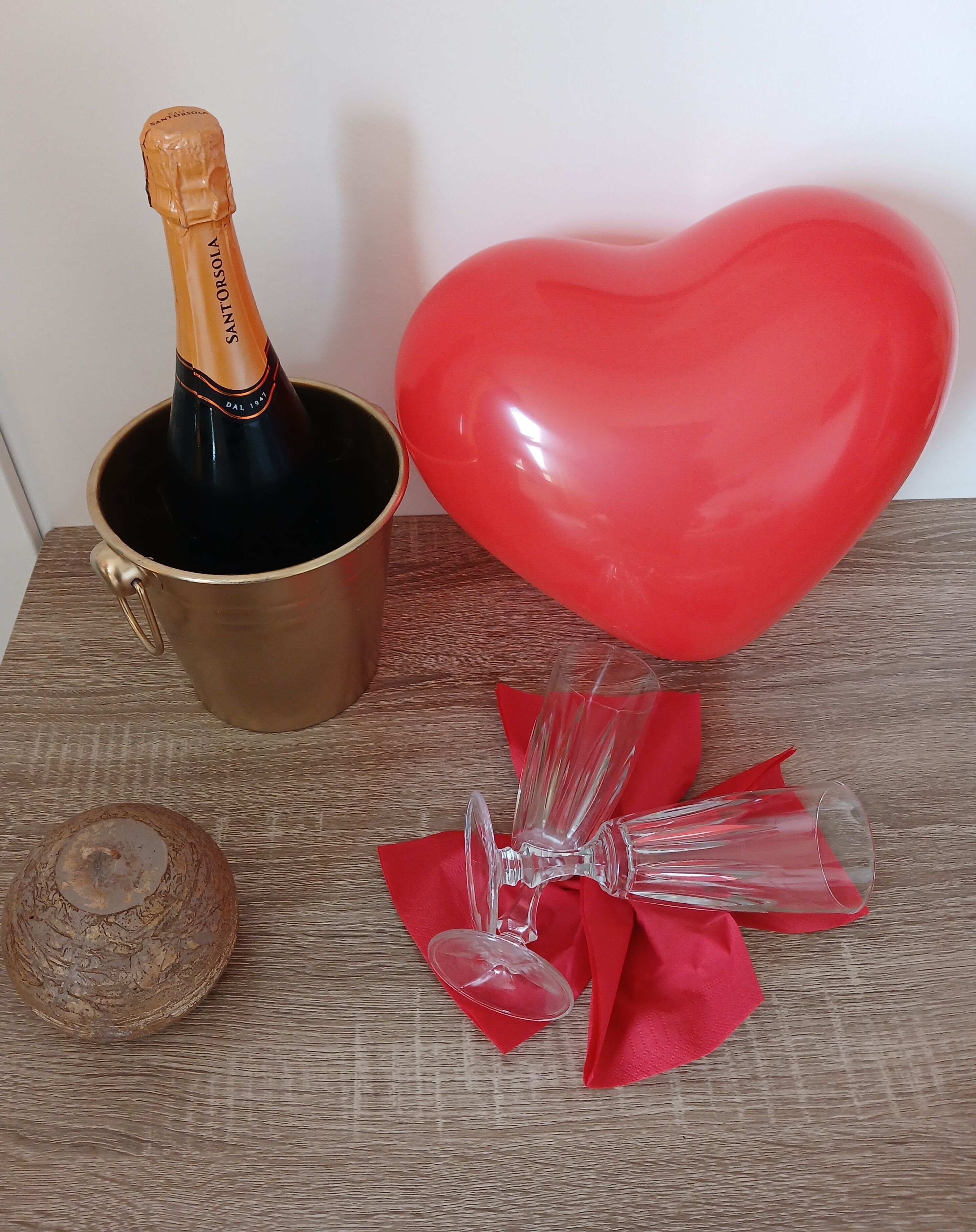Une bouteille de champagne dans un seau doré, un ballon rouge en forme de cœur et un verre avec une serviette rouge.