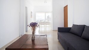 A modern living room with a dark grey sofa, a wooden coffee table with a vase of dried flowers, and a view of a balcony with a white railing. - Neos Kosmos 1Bd Apt with Stunning Views (Athens)