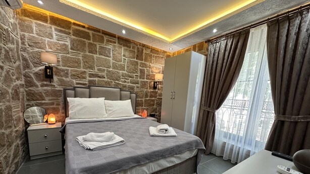 Ein modernes Schlafzimmer mit Steinwand, einem Bett mit grauer Tagesdecke, Nachttischlampen, einem Kleiderschrank und Blick durch Vorhänge.