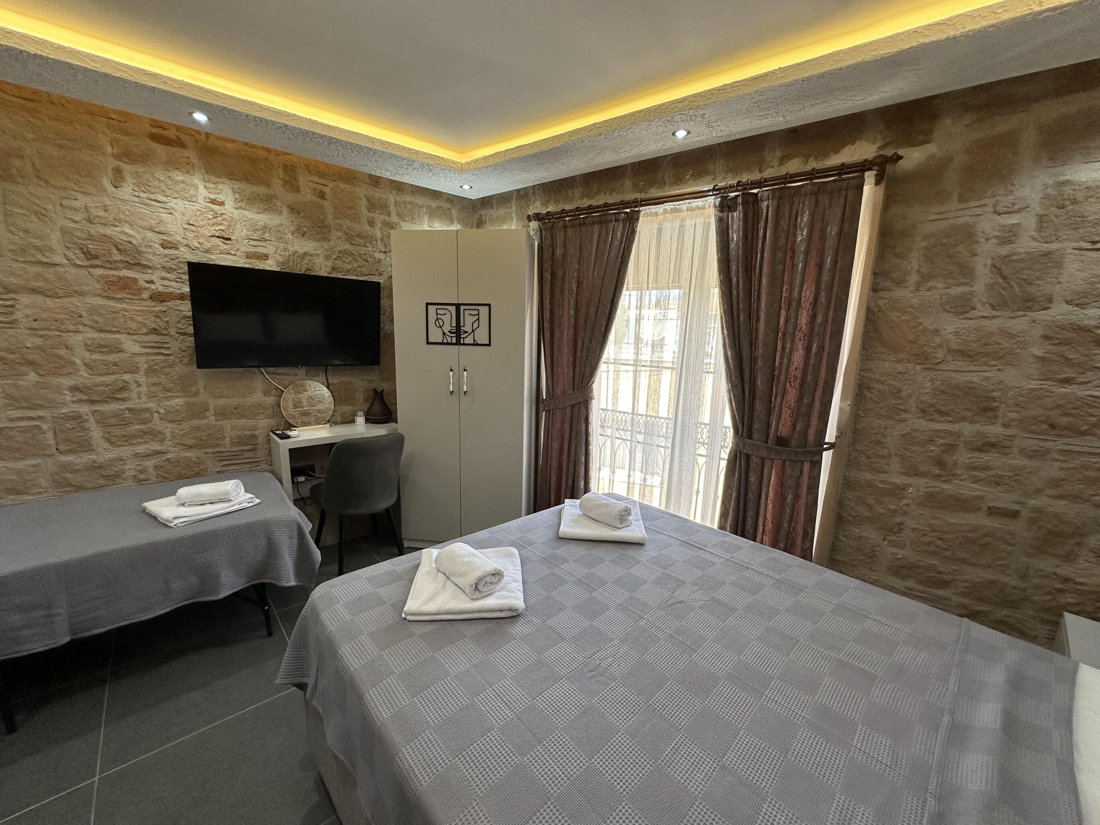 A hotel room with two beds, a desk, a TV, and stone walls.