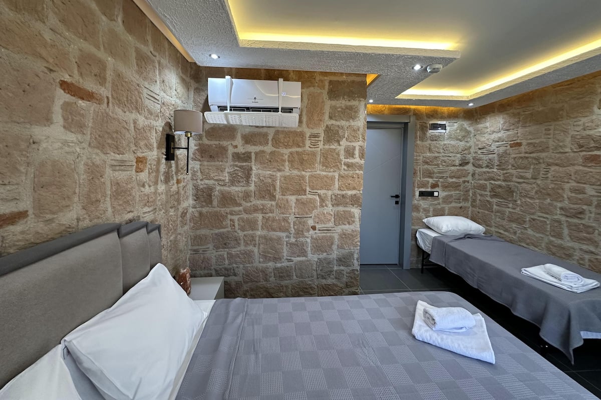 Quarto com paredes de pedra, uma cama com cabeceira, duas mesas de cabeceira, uma luminária e um ar condicionado, além de uma porta.