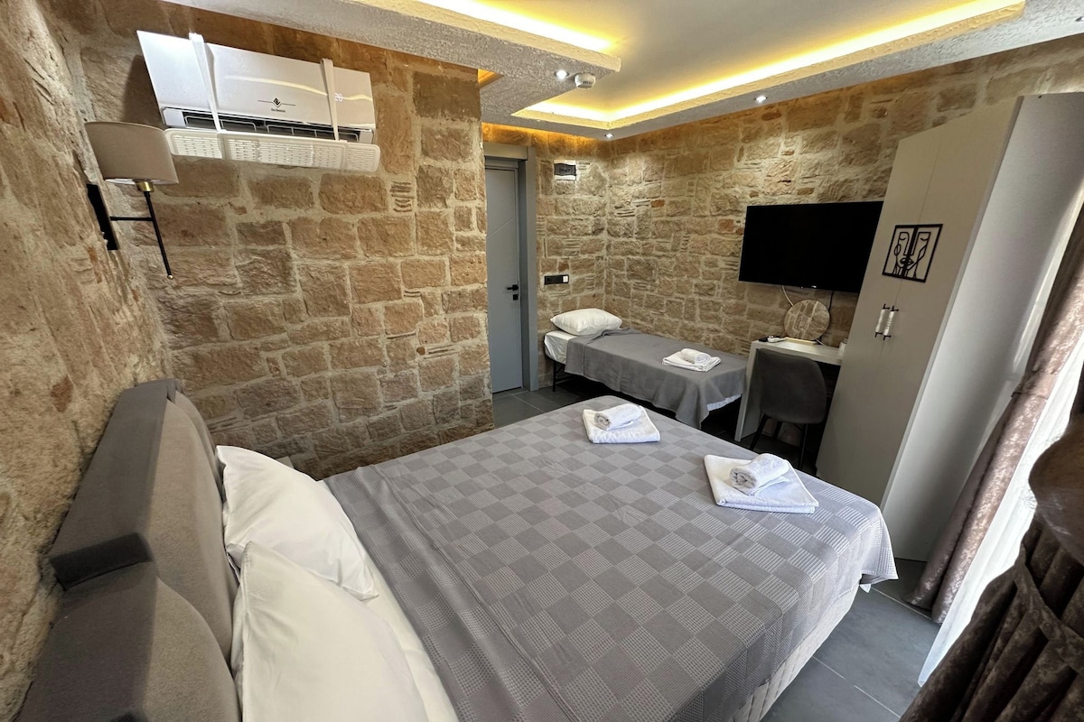 A hotel room with a stone wall, a bed with a grey checkered bedspread, a desk with a chair, and a flat-screen TV mounted on the wall.