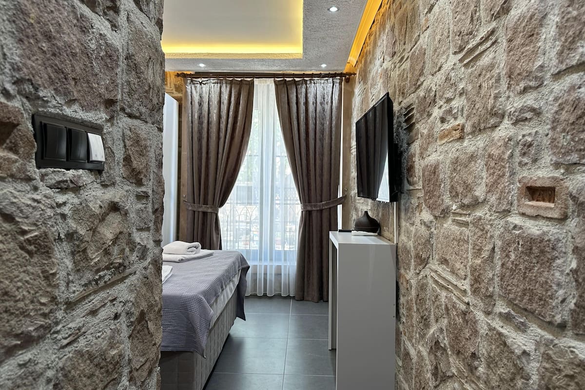 Quarto com paredes de pedra, uma cama, mesa de cabeceira, televisão e janela com cortinas.