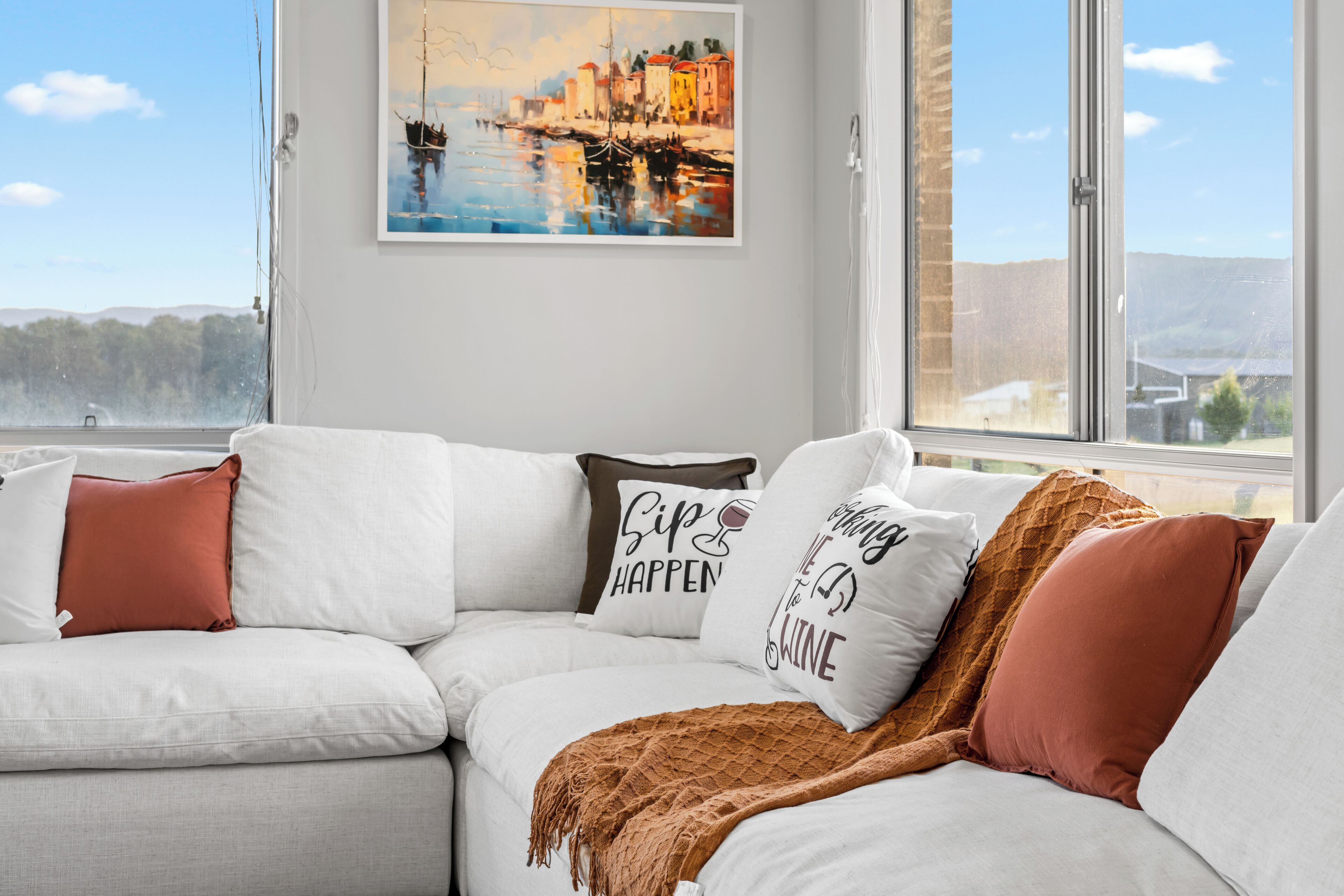 A modern living room with a white sofa, a painting of a coastal scene, and a view of the outdoors.