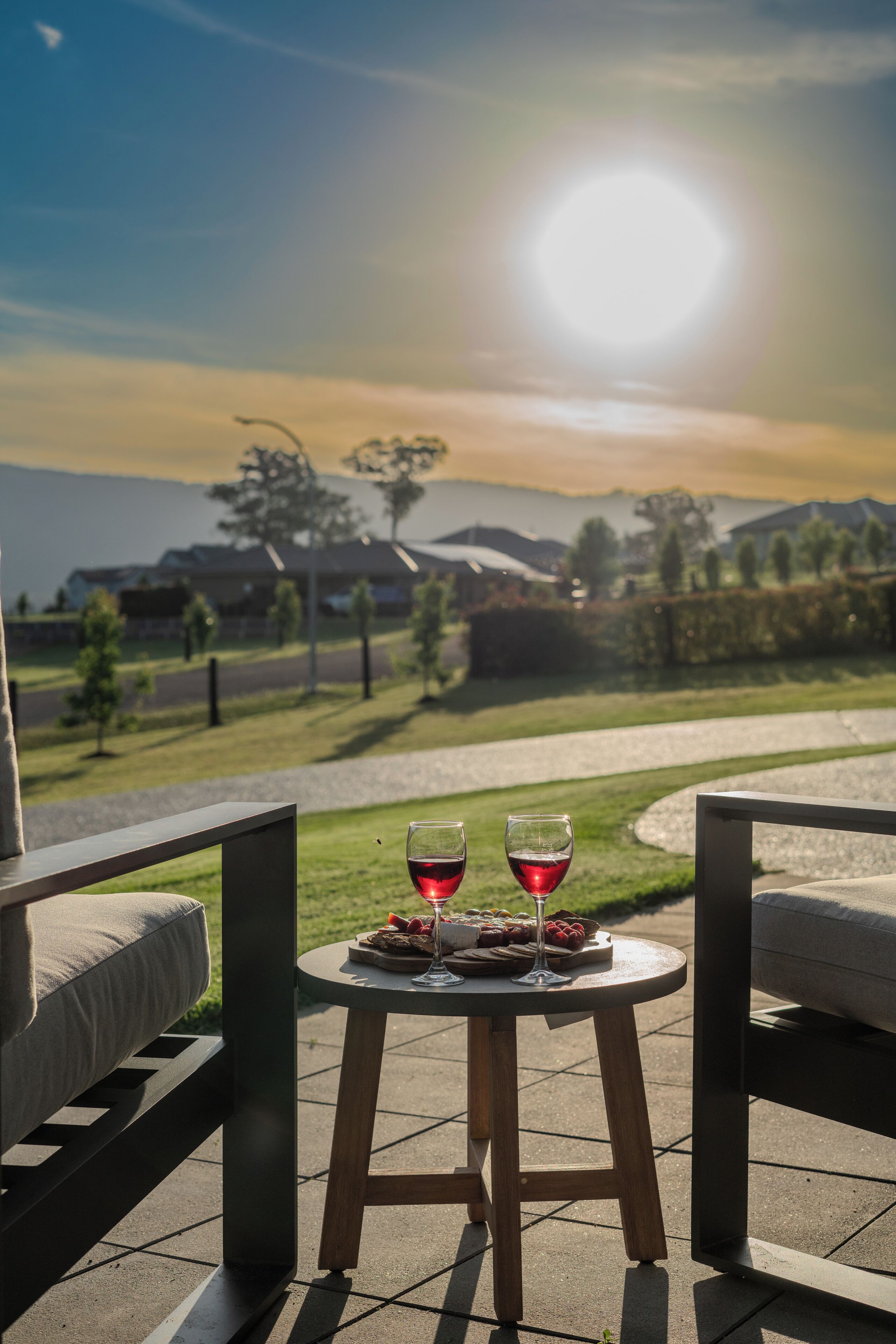Een terras met twee stoelen, een klein rond tafeltje met twee glazen rode wijn en een bord eten, en een mooi uitzicht op een weg en huizen in de verte.