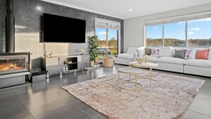 A modern living room with a fireplace, a flat-screen TV, a white sofa, and large windows offering a view of the outdoors. - Serenity Estate Hunter Valley (Millfield)
