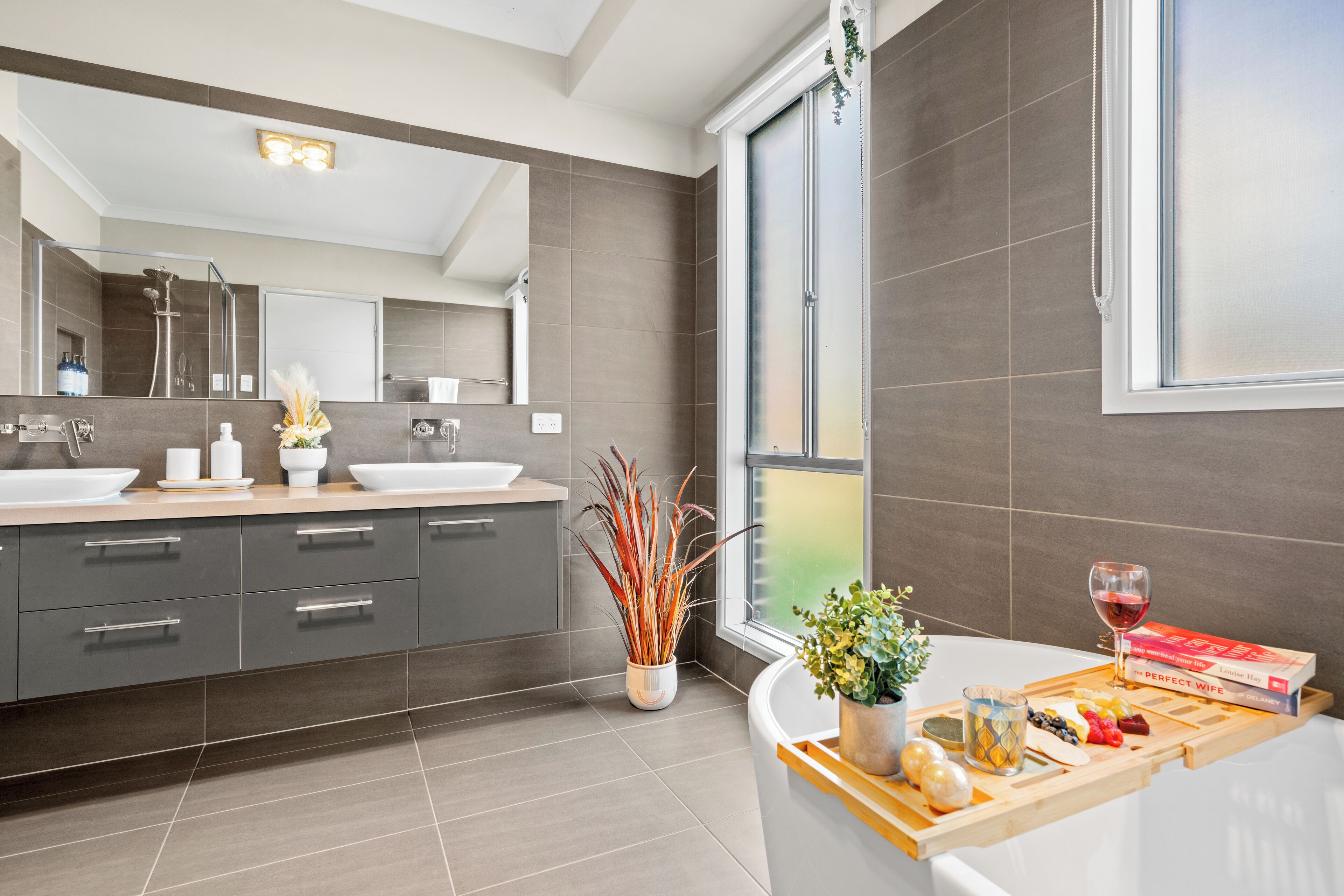 Un baño moderno con un espejo grande, dos lavamanos y una ducha. En la encimera hay una bandeja con frutas, una copa de vino y un libro.