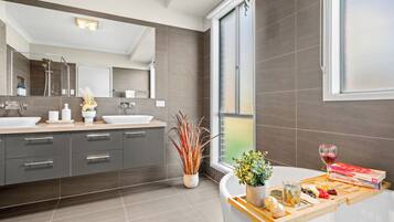 Un baño moderno con un espejo grande, dos lavamanos y una ducha. En la encimera hay una bandeja con frutas, una copa de vino y un libro.