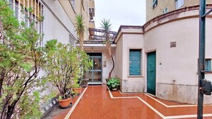 Une ruelle étroite bordée de plantes en pot et donnant sur l’entrée d’un immeuble.